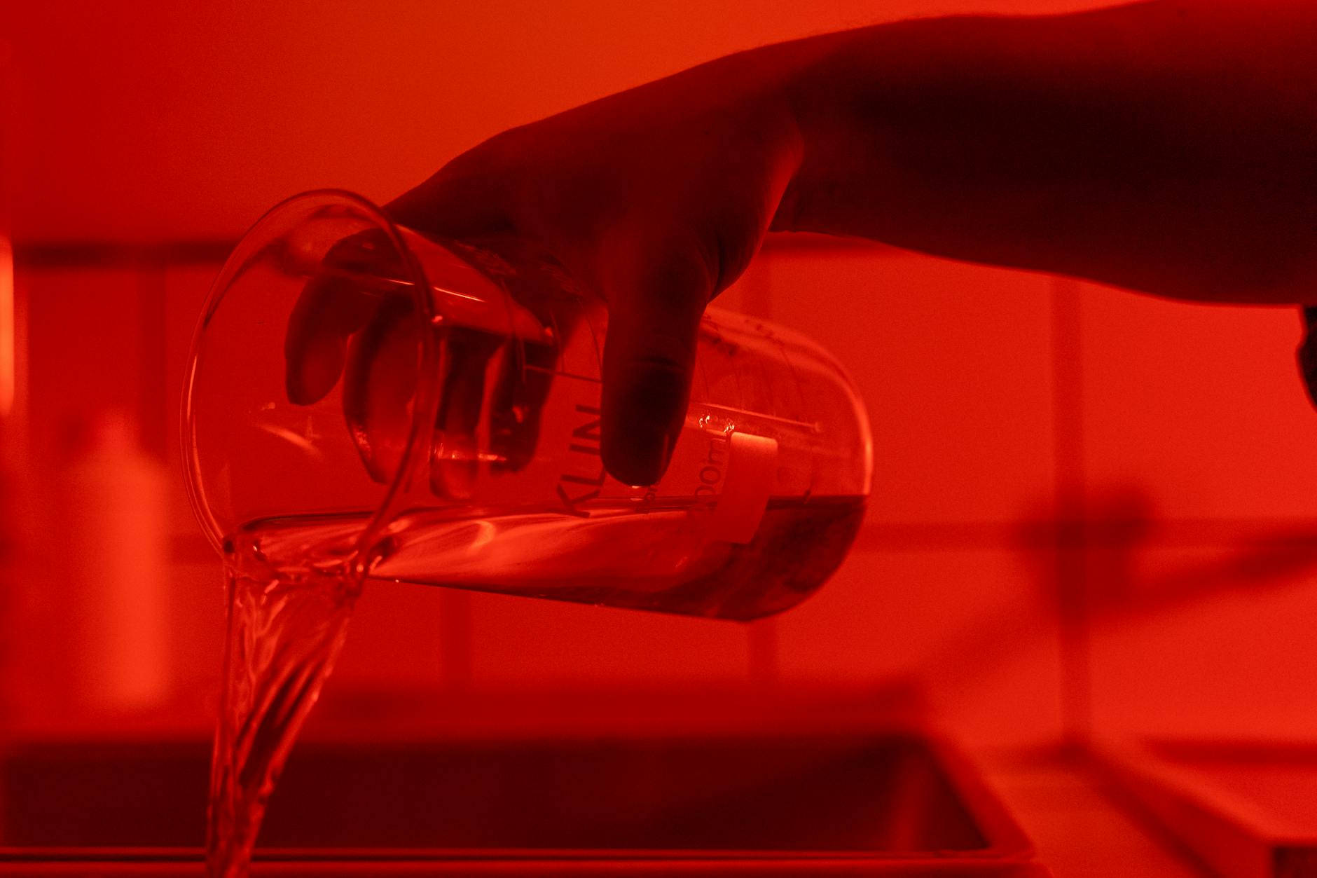 A hand pouring liquid from a beaker under atmospheric red lighting indoors.