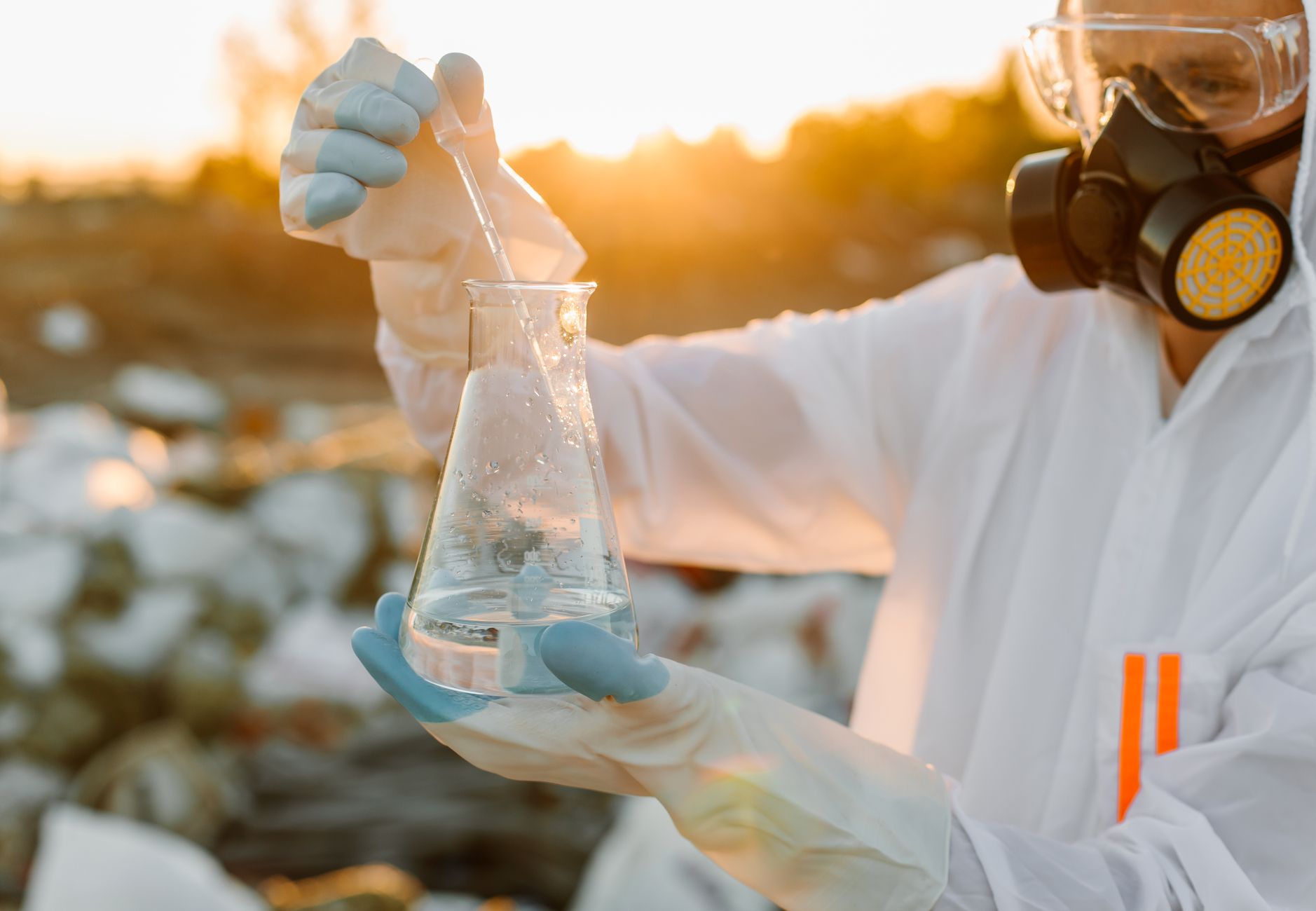 Researcher in protective suit with Erlenmeyer flask outdoors at sunset.