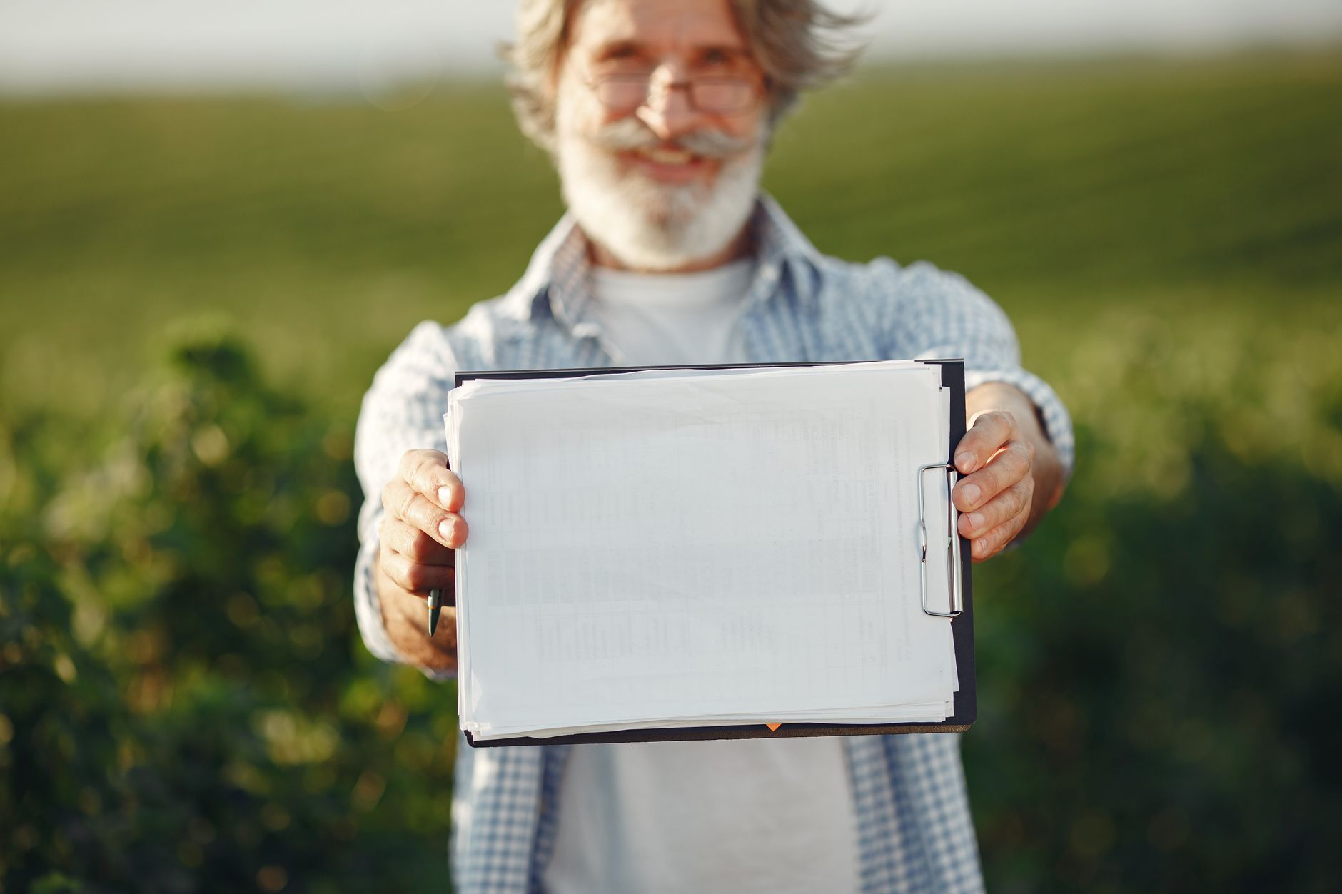 Bearded senior man with glasses holding a clipboard in a green field on a sunny day.