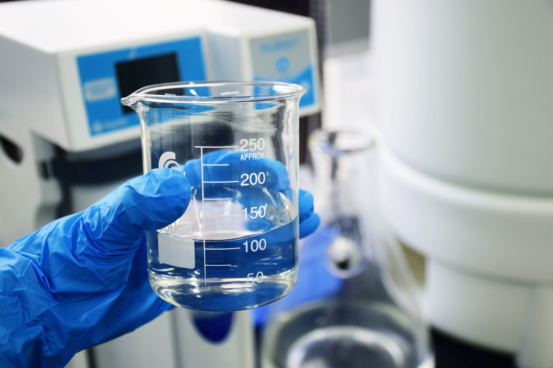 Gloved hand holding a beaker with clear liquid, set against a laboratory background.