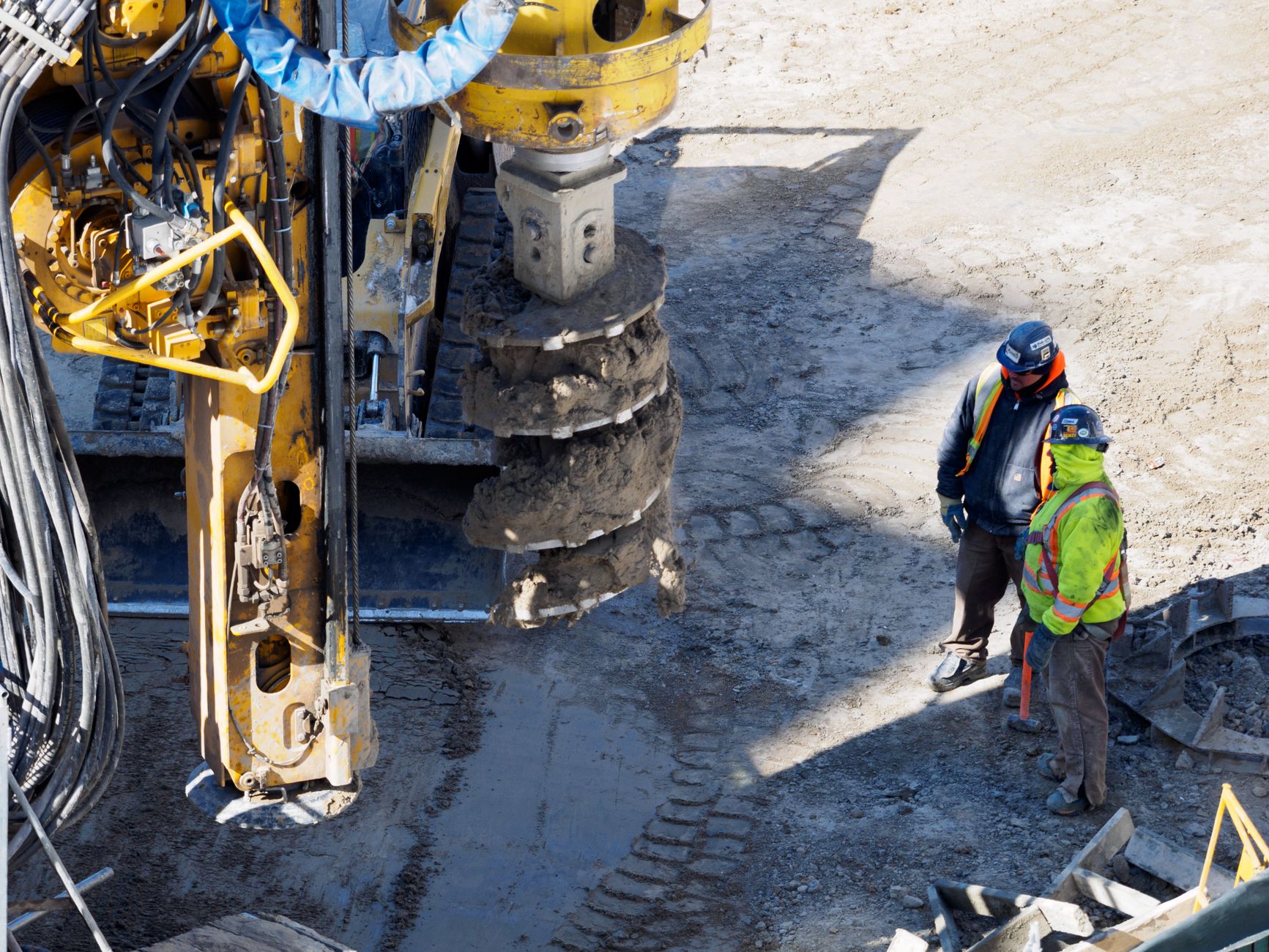 Two workers in protective gear at a construction site with heavy drilling machinery.