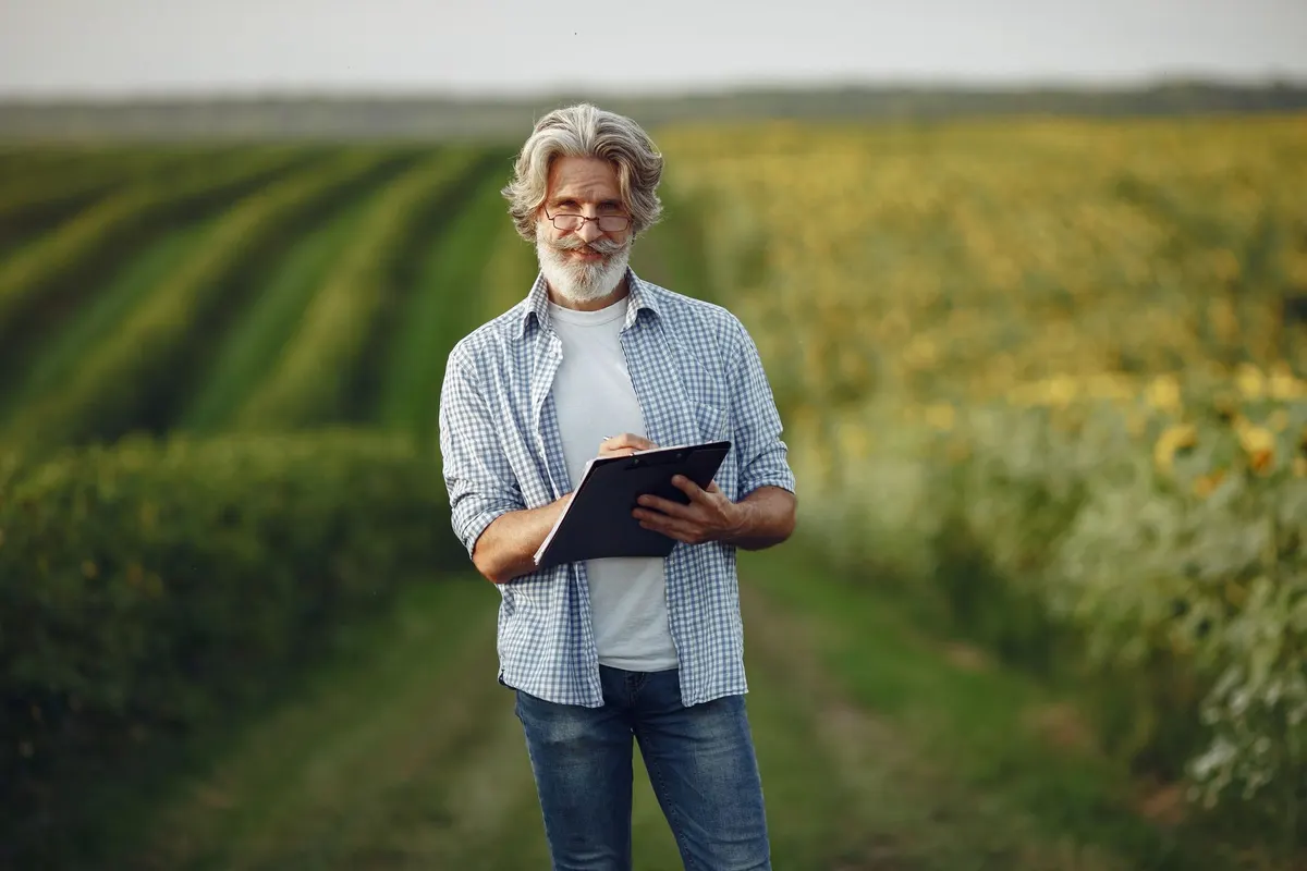 Senior farmer taking notes in sunny countryside field, wearing eyeglasses.
