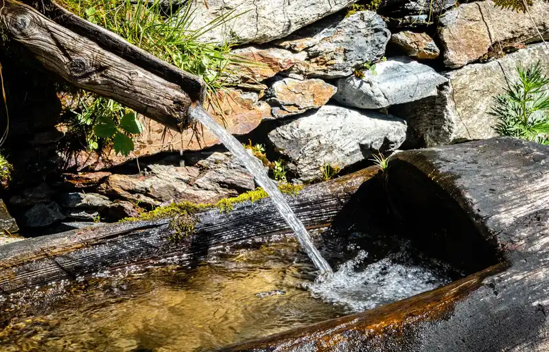 Clear water flows from a rustic wooden spout into a basin, set against a stone wall.